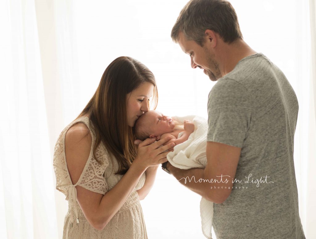 mom and dad kissing newborn baby's head next to window