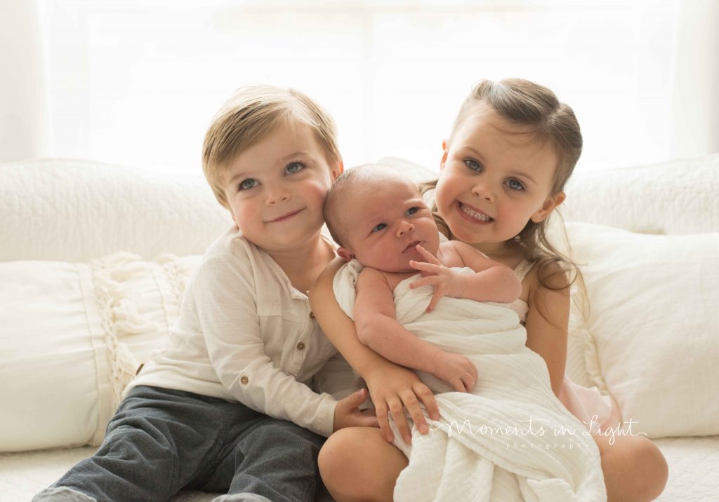 siblings holding newborn baby on bed smiling