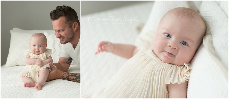 A father helps his small baby girl sit up on a white bed. 