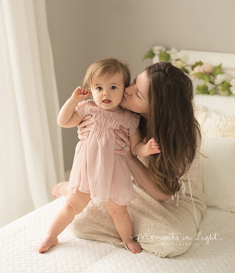 A baby girl stands up on a bed while her mother supports her from behind. 