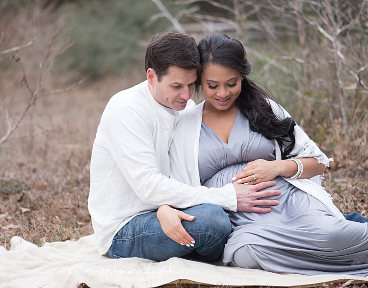 An expecting couple sits on a white blanket. 