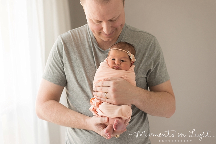 A baby swaddled in a pink blanket is held in front of her father. 