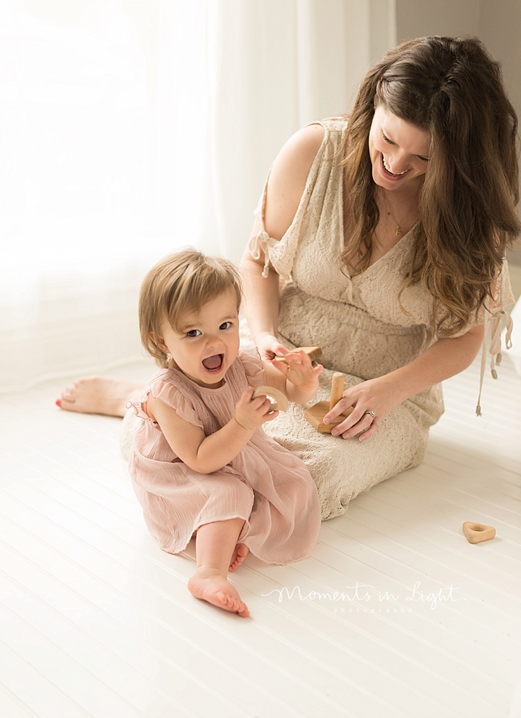 A baby plays with wooden toys on a white floor with her mom nearby. 