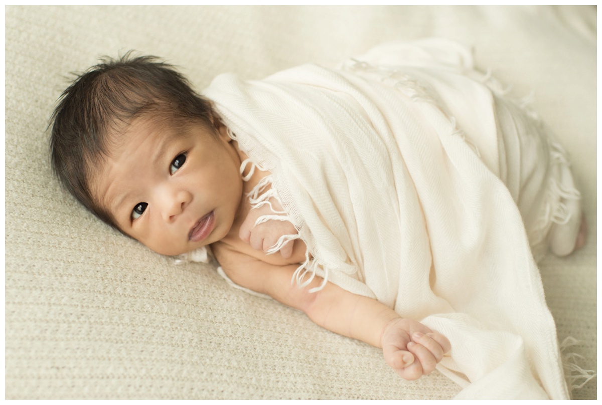 Baby swaddled in a white wrap looking into the camera by a Houston newborn photographer