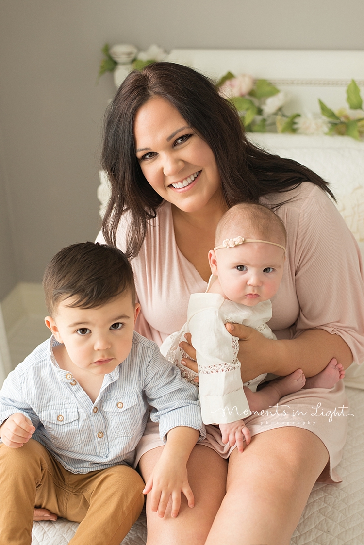 Two young children sit in their mother's lap on a white bed. 