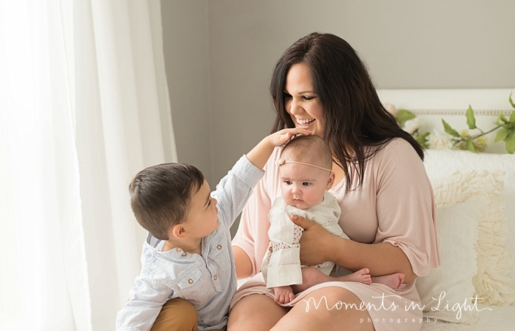 A mother wearing a pink dress sits on a bed with her babies. 