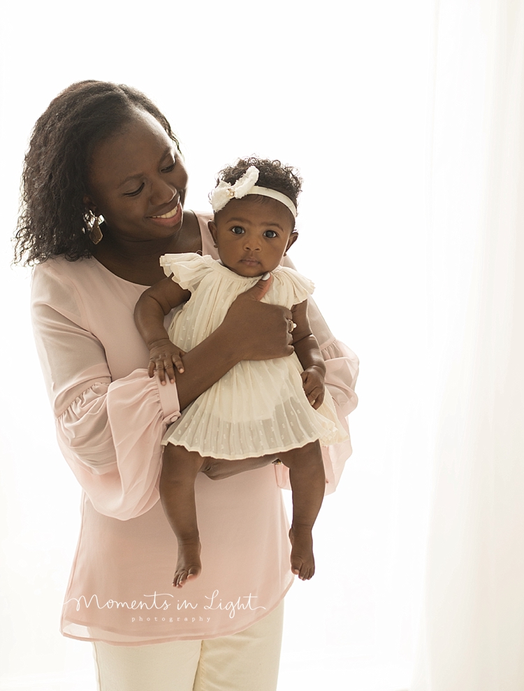 A baby wearing a white dress is held by her mother in a bright room. 