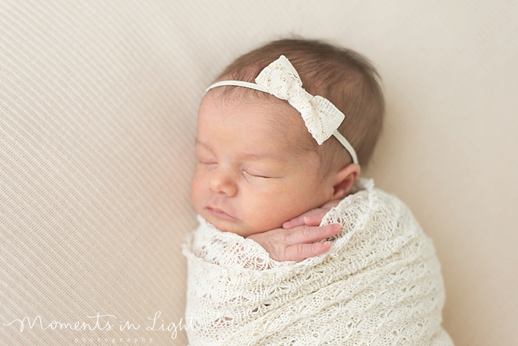 A newborn swaddled in white and wearing a white bow take a a nap. 