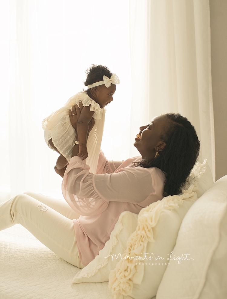 A mom sits on a bed and holds her baby up in the air. 