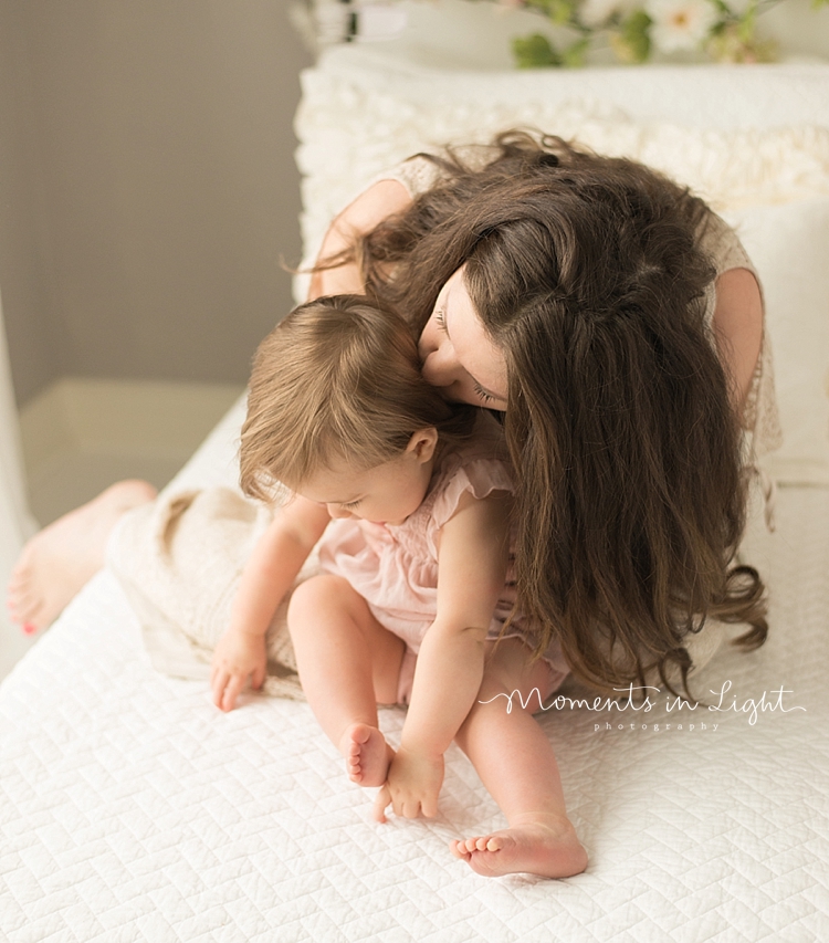 A mother bends over to kiss her baby on the neck while she tickles her. 