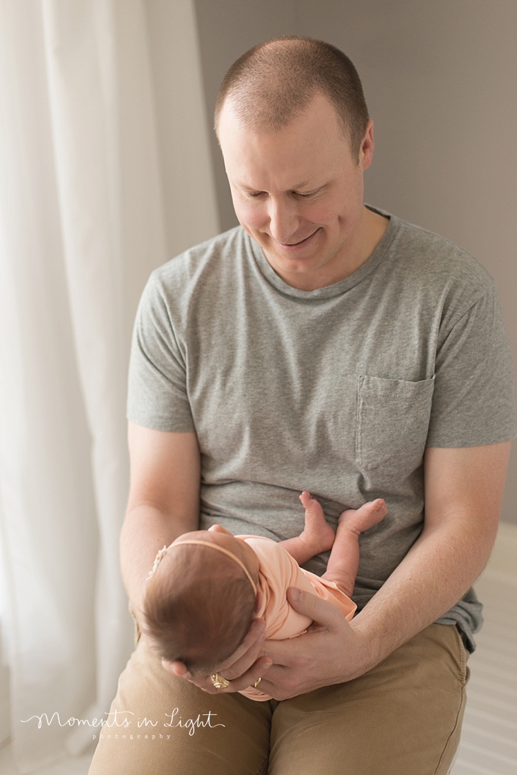 A father sits and holds his newborn in front of him on his lap. 