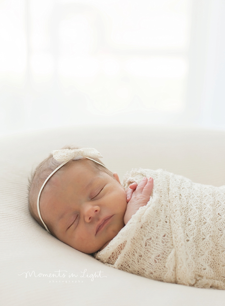 A newborn baby girl naps in a white room while wrapped in white. 