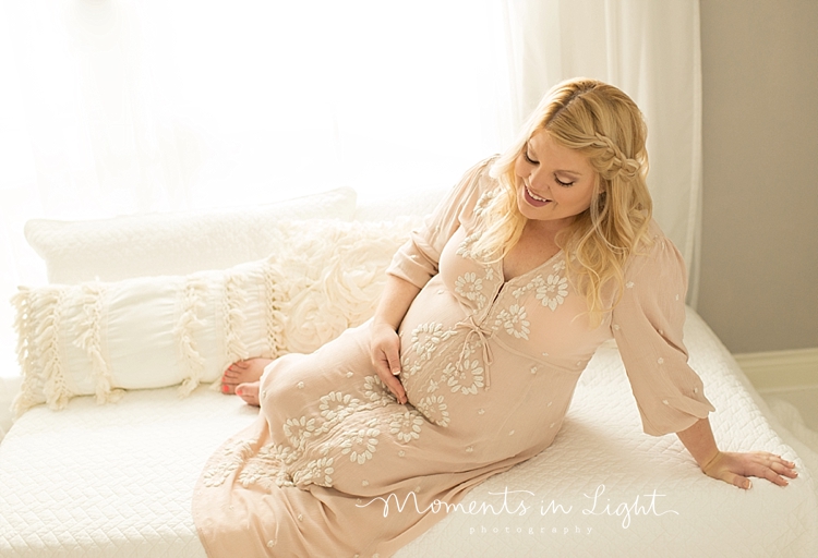 A pregnant mom wears a white dress and sits on a bed. 