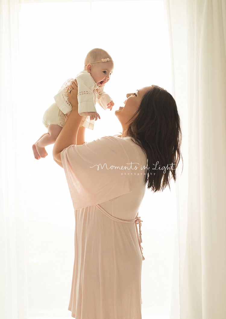 Standing next to a bright window, a mother holds her baby up high. 