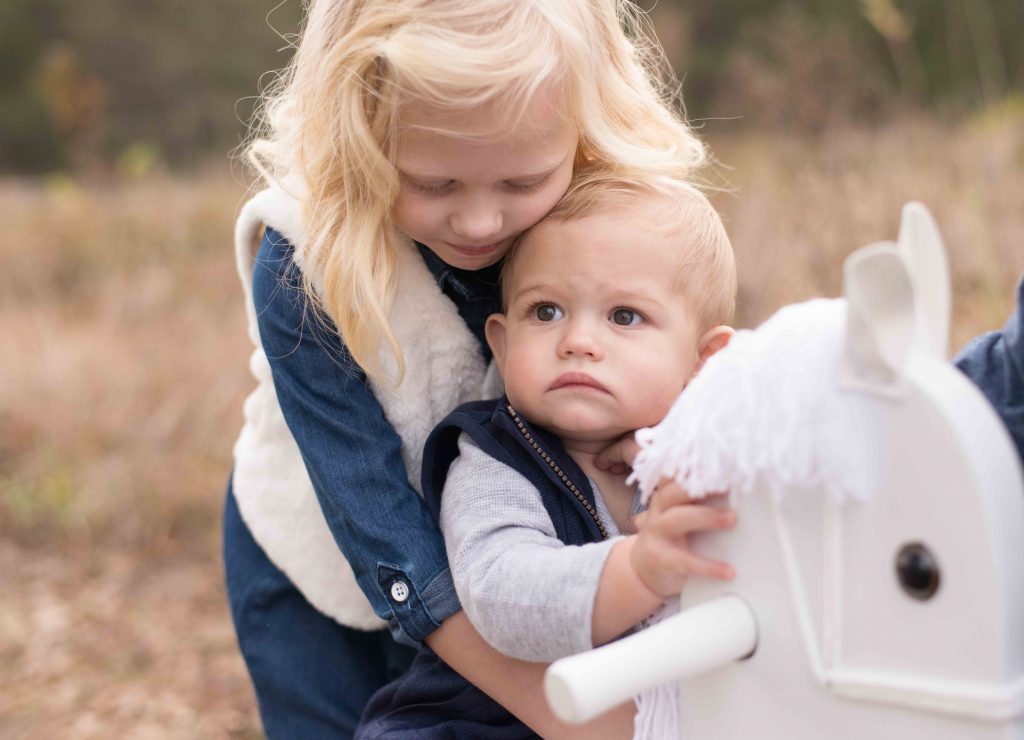 baby boy on a rocking horse being held by big sister 