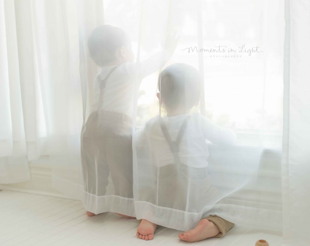 twin little boys playing in white curtains by window in The Woodlands, TX